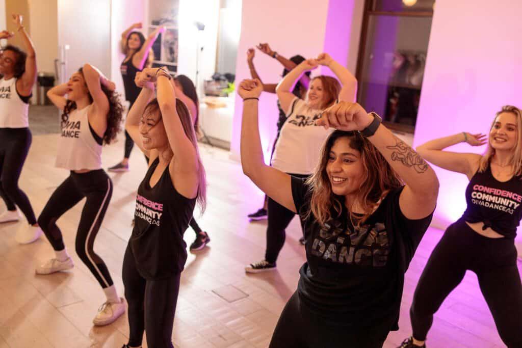A group of women at a DivaDance class, smiling and holding their arms up. The room has wooden flooring and is lit with pink light. This is one of the best bachelorette party ideas in Charlotte!