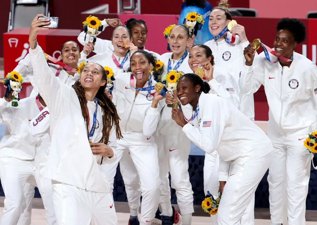A group of female athletes in white uniforms pose for a selfie, showing their gold medals and holding bouquets on a podium.
