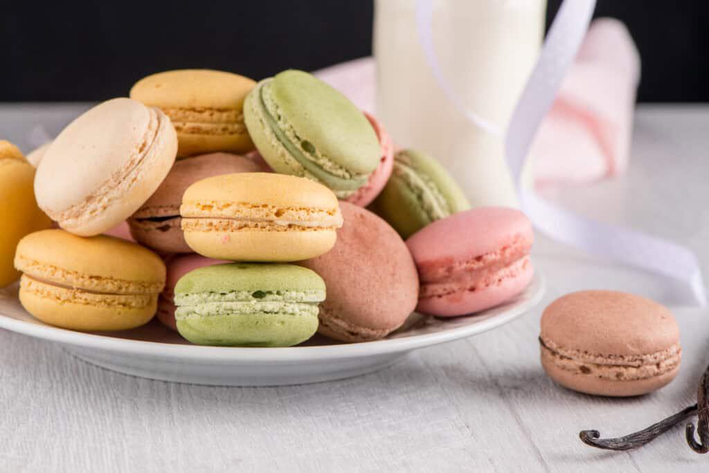 A plate of assorted colorful macarons is placed on a light-colored surface next to a glass of milk.