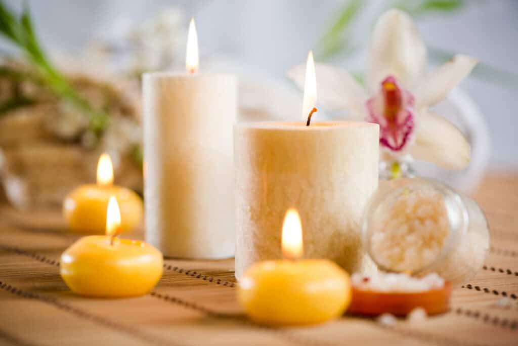 Four lit candles of varying sizes are arranged on a mat with white flowers and a small open container of bath salts in the background.