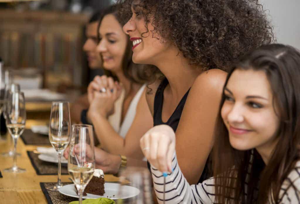 Four women sit at a dinner table, smiling and engaging in conversation. Wine glasses and dessert plates are on the table in front of them—one of the many delightful bachelorette party ideas in St. Augustine.