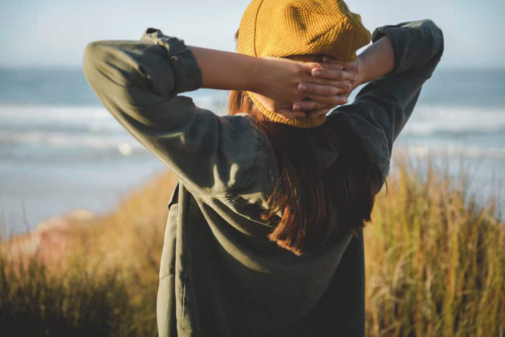 A person wearing a yellow hat and a green jacket stands facing the ocean with their hands on the back of their head.