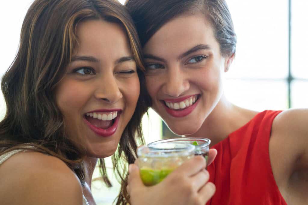 Two women smiling and holding a drink together, with one woman winking at the camera.