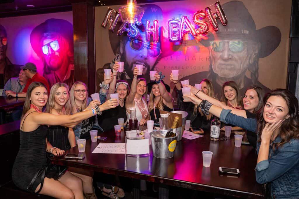 A group of people at a bar celebrate by raising their drinks at a lively Nashville bachelorette party. They are seated at a long table with a bucket of ice and drinks in the center. "NASH BASH" is spelled out in balloons above them, adding to the festive atmosphere.