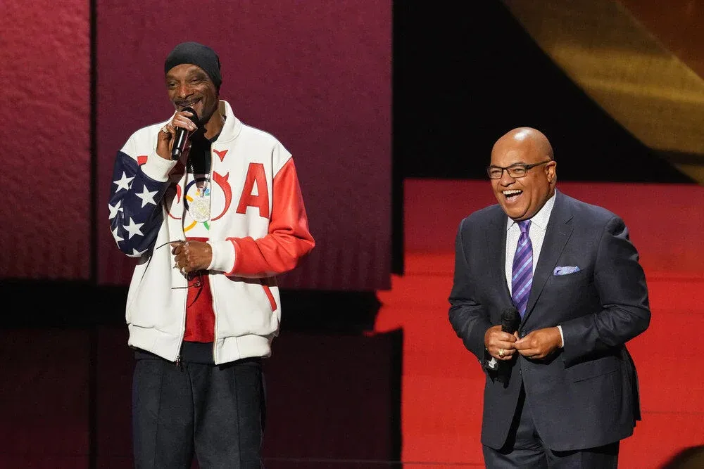 Two men are on stage. The man on the left is holding a microphone, wearing a USA jacket, and the man on the right, in a suit, is smiling.