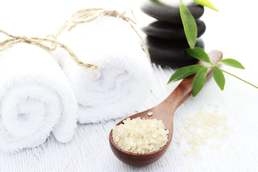 Two rolled white towels, a wooden spoon with bath salts, and black massage stones with green leaves on a white surface.