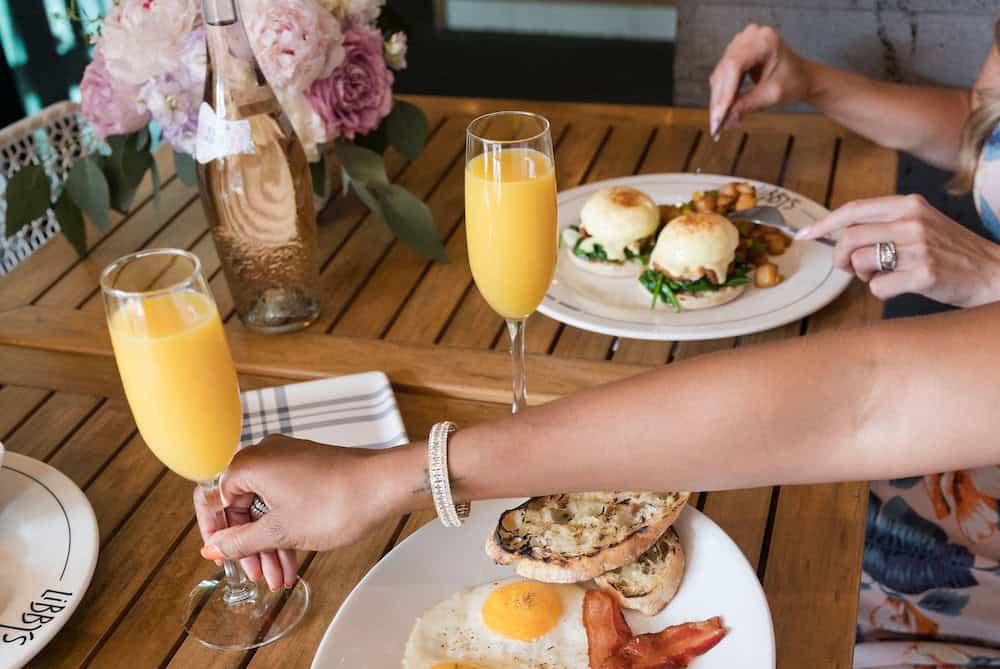 Two people enjoying brunch with plates of eggs, bacon, bread, and potatoes on a wooden table. The table also has two glasses of orange juice and a vase of flowers.