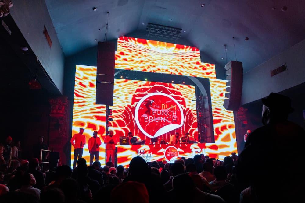 A vibrant event with a crowd watching a performance on a stage featuring a bold "the rum punch brunch" logo.