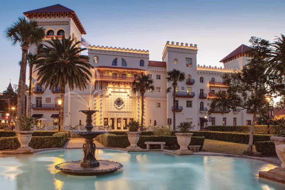 A grand white building with turreted features stands behind a fountain in a courtyard surrounded by palm trees and neatly trimmed hedges. Ideal for bachelorette party ideas in St. Augustine, the warm lighting enhances the hotel's entrance.