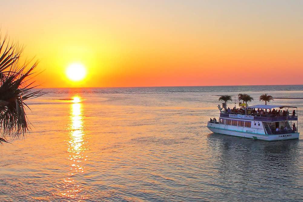 A boat carrying passengers sails on a calm body of water at sunset. The sky is orange and the sun is low on the horizon. Palm trees are visible on the left and on top of the boat.