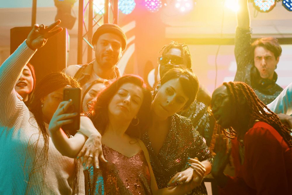 A group of people, illuminated by colorful lights, gather closely together, some posing for a selfie while others strike various expressive poses.