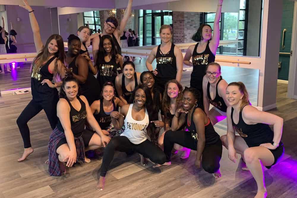 A group of women in at DivaDance, posing together and smiling, dressed in workout attire—one of the top 8 bachelorette party ideas in Tallahassee. The room has large mirrors, wooden floors, and large windows.