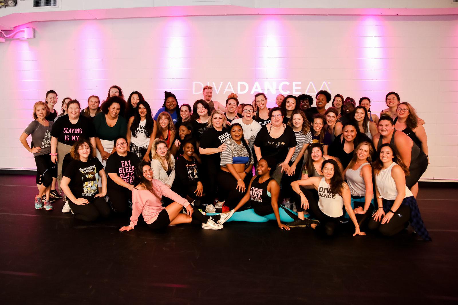 A large, diverse group of DivaDance members of all ages, sizes, and backgrounds smile together for a photo in front of the pink-lit DivaDance logo after class