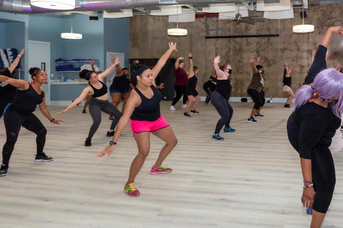 A full DivaDance class moves in unison with arms raised, led by a member in a black tank and pink shorts, in a bright modern studio