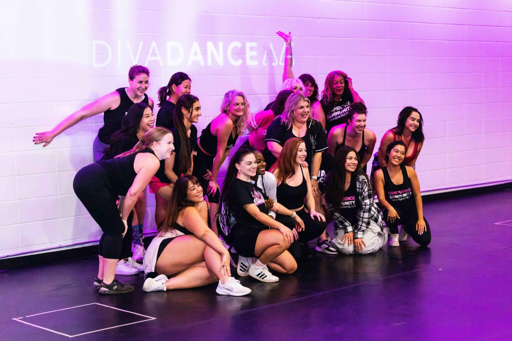 Sixteen women from a DivaDance class crouch and kneel together, smiling for a group photo against a white studio wall with the DivaDance logo projected in purple light.