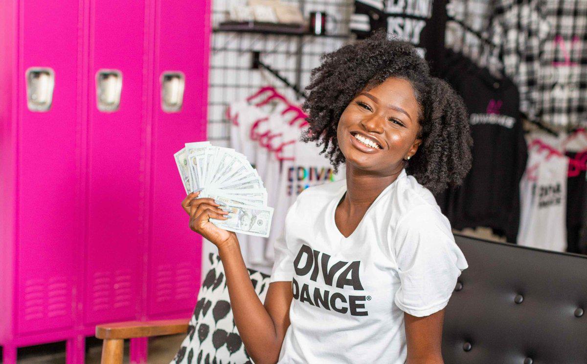 A smiling woman in a DivaDance T-shirt holds a fanned stack of cash in front of bright pink lockers and a studio merchandise wall.