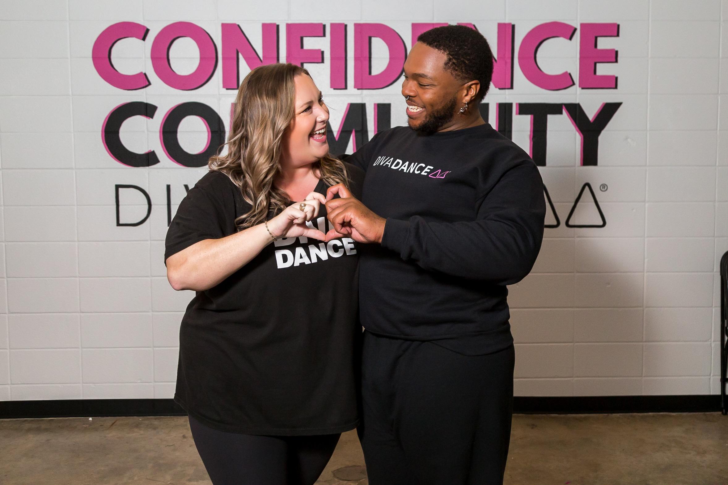 Two laughing DivaDance members forming a heart shape with their hands together, standing in front of a Confidence Community wall mural