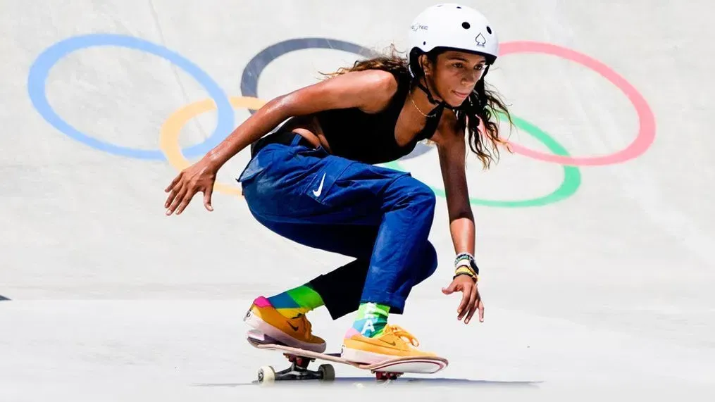 A person wearing a helmet and protective gear rides a skateboard in a skatepark with Olympic rings visible in the background.