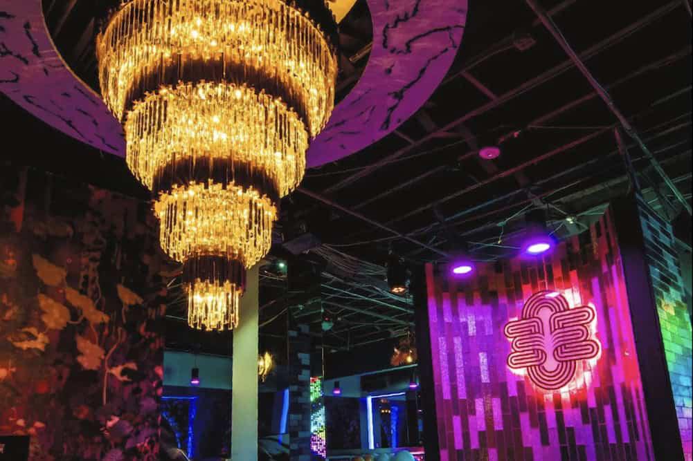 A lavish chandelier hangs from the ceiling in a dimly lit room with a neon double "E" sign on a wall.