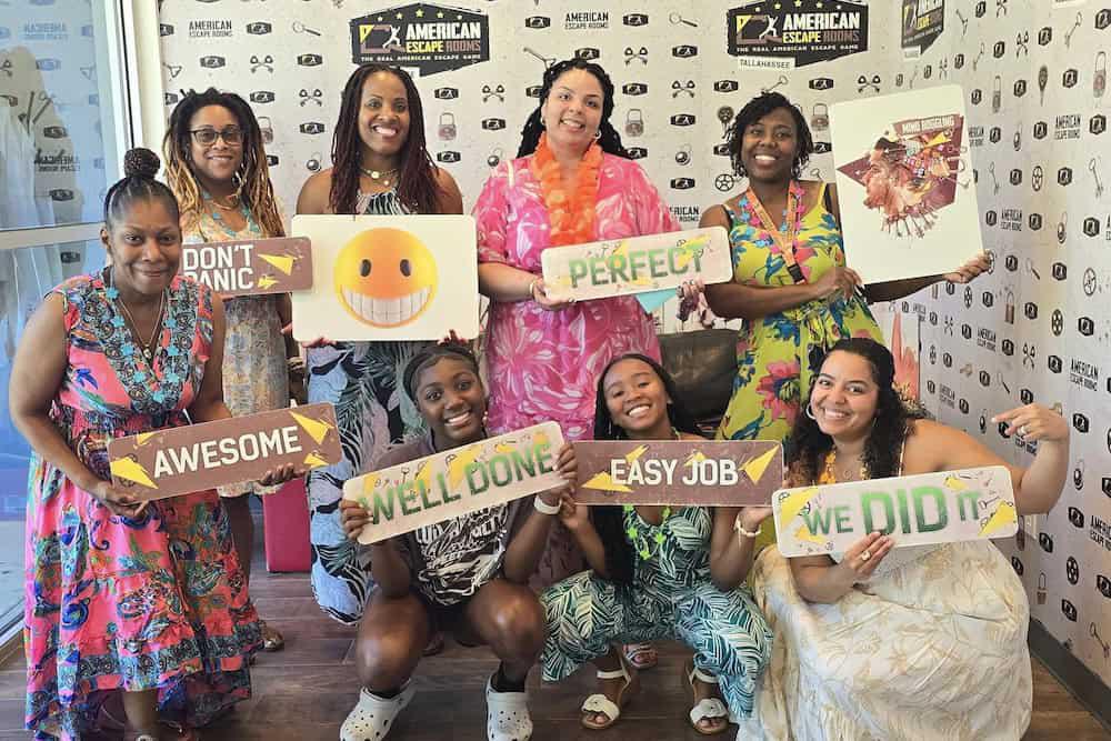 A group of people posing with celebratory signs reading "Don't Panic," "Awesome," "Well Done," "Perfect," "Easy Job," and "We Did It" in front of an escape room backdrop, undoubtedly one of the Top 8 Bachelorette Party Ideas in Tallahassee.