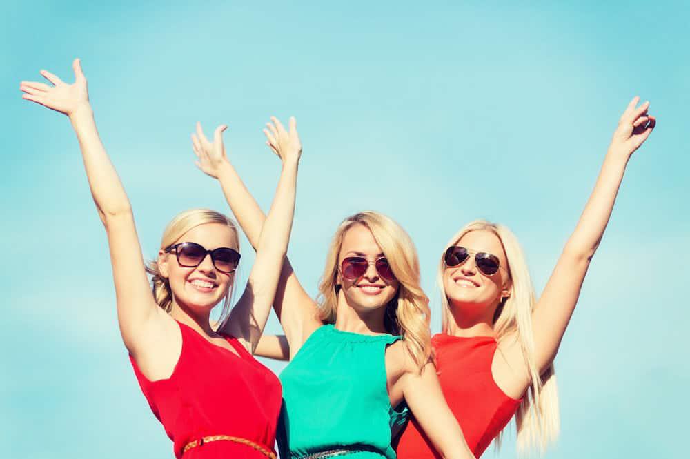 Three joyful women wearing sunglasses and colorful dresses raise their arms against a clear blue sky.