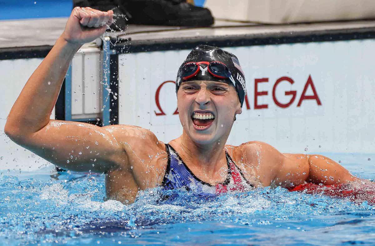 A swimmer celebrates with a fist pump in the pool, wearing a swim cap and goggles, standing near a wall with an Omega logo.