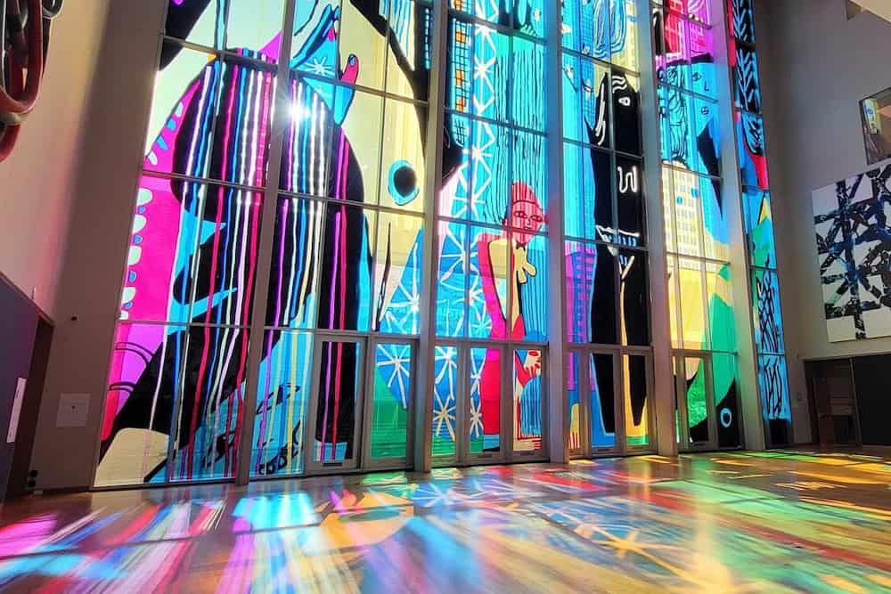 Brightly lit stained glass windows with abstract designs cast colorful reflections on a wooden floor in an art gallery.