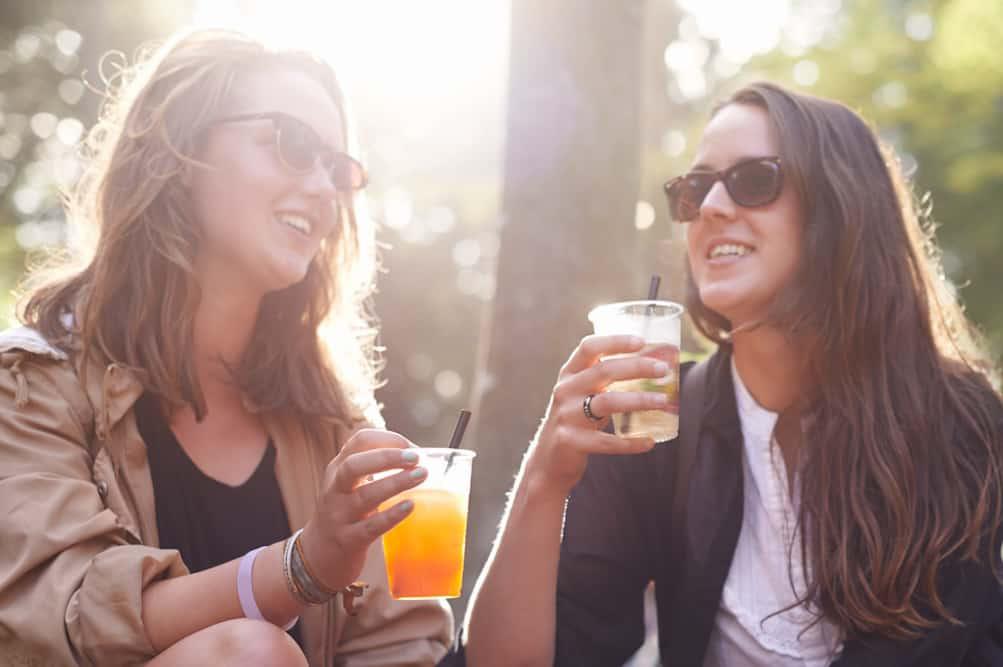 Two women laughing and enjoying drinks outdoors in sunlit scene.