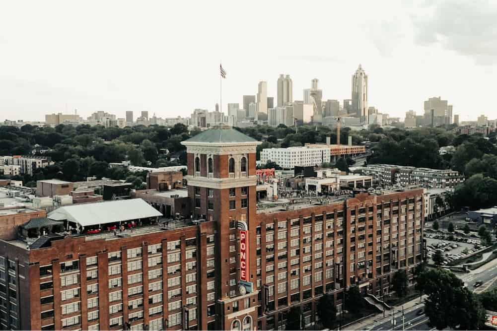 Aerial view of a historic brick building with a distinctive tower in the foreground, with a backdrop of a sprawling city skyline under a cloudy sky.
