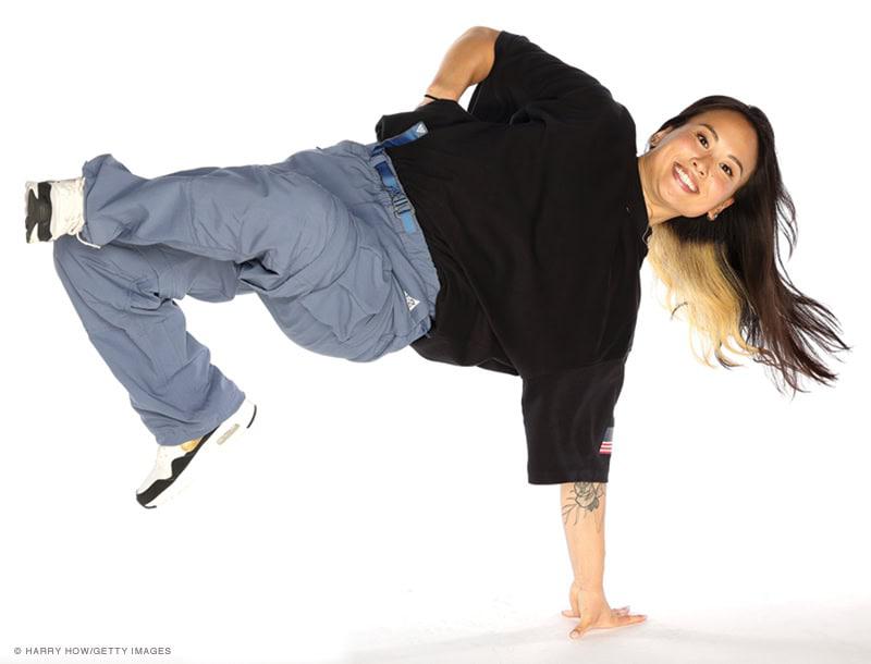 Person performing a breakdance move, balancing on one hand with legs extended. They are wearing a black shirt, blue pants, and sneakers, and have long hair with a streak of blonde.