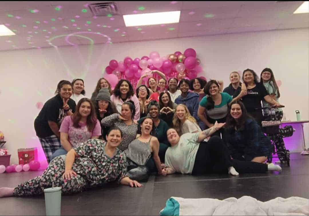A group of people posing for a photo in front of pink and white balloon decorations. Some are sitting on the floor, while others are standing behind them. The room has a relaxed atmosphere.