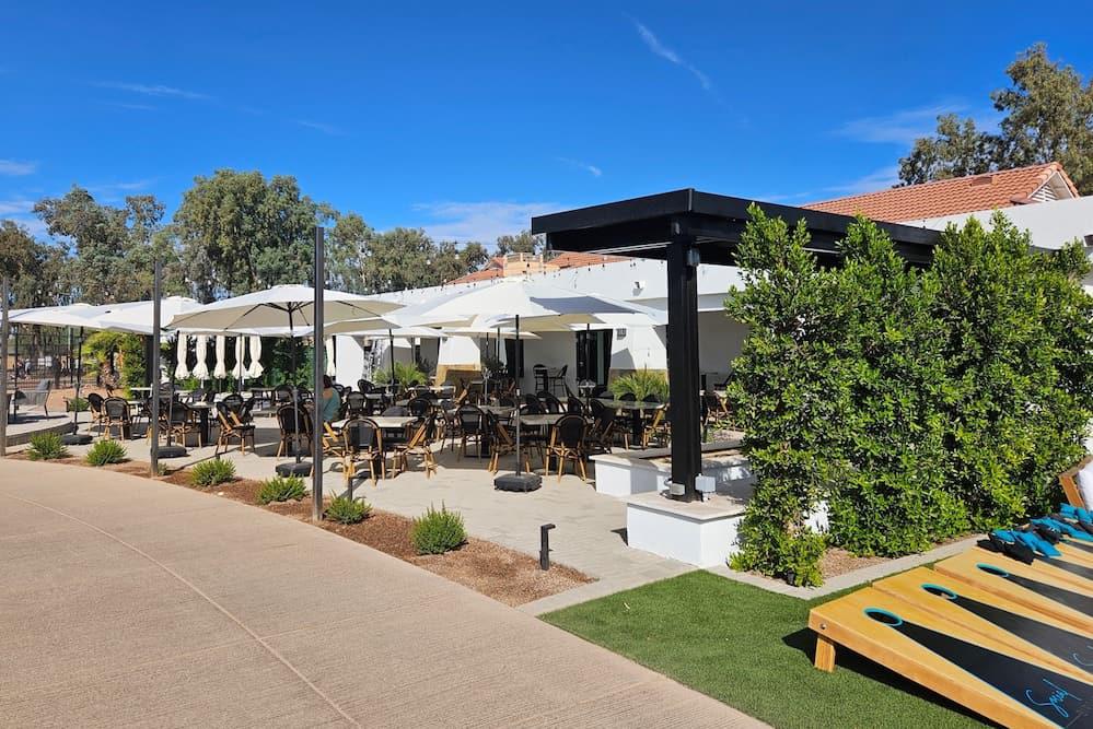 Outdoor dining area with shaded seating, greenery, and a games zone under a clear blue sky.