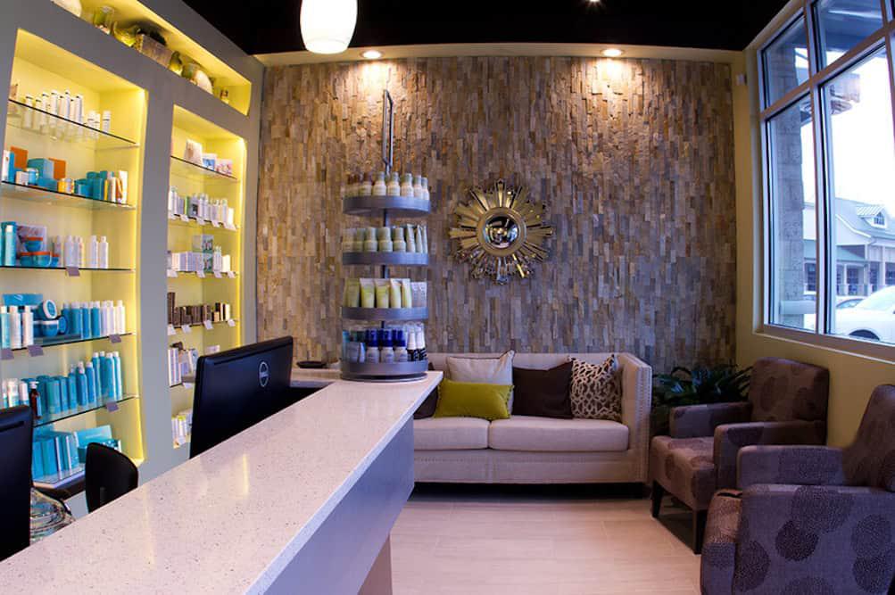 Modern salon interior with a reception desk, product shelves, beige couch, and two chairs. Decor includes a textured wall, sunburst mirror, and soft lighting—perfect for pampering before exploring the top 8 bachelorette party ideas in Tallahassee.