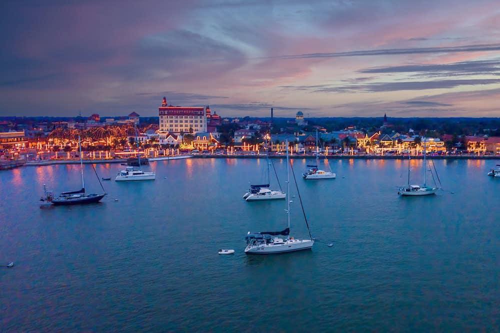 Evening view of a coastal town with boats anchored in the harbor and buildings along the waterfront illuminated by lights.