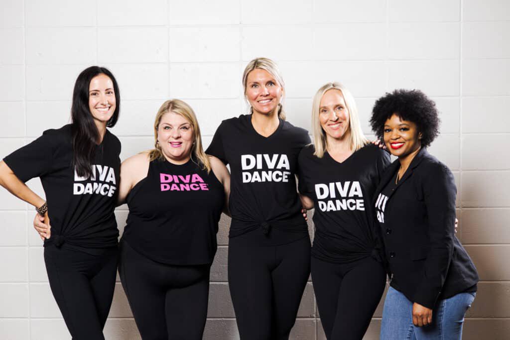 Five women standing side by side, all wearing black "Diva Dance" shirts, smiling in front of a white brick wall.
