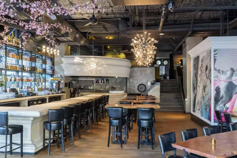 A modern restaurant interior features a long bar with stools, tables and chairs, artistic wall decor, and hanging light fixtures. Stairs in the background lead to an upper level.