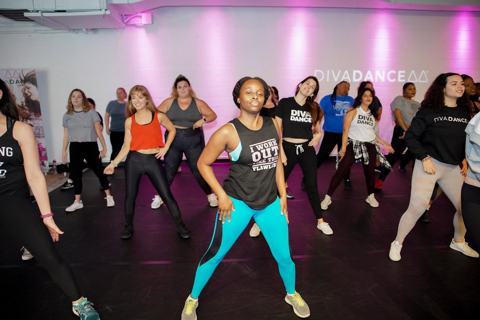 A DivaDance member in a tank top and bright leggings stands with one hand on her hip during class, with a diverse group of dancers behind her and the DivaDance logo on the wall