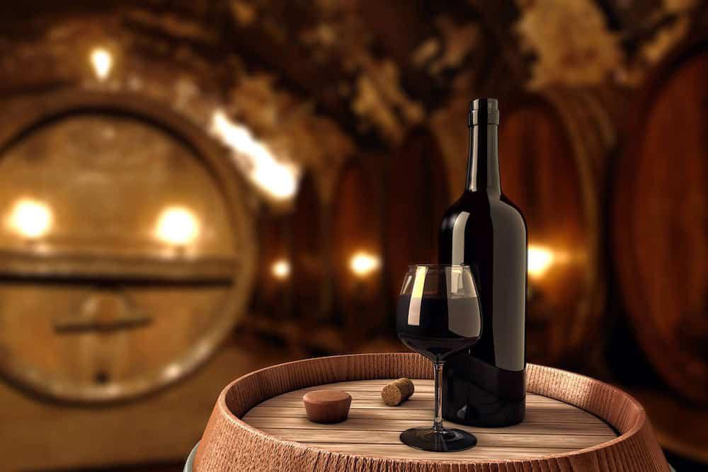 A wine bottle and a filled wine glass sit on a wooden barrel in a dimly lit wine cellar with large wooden casks in the background.
