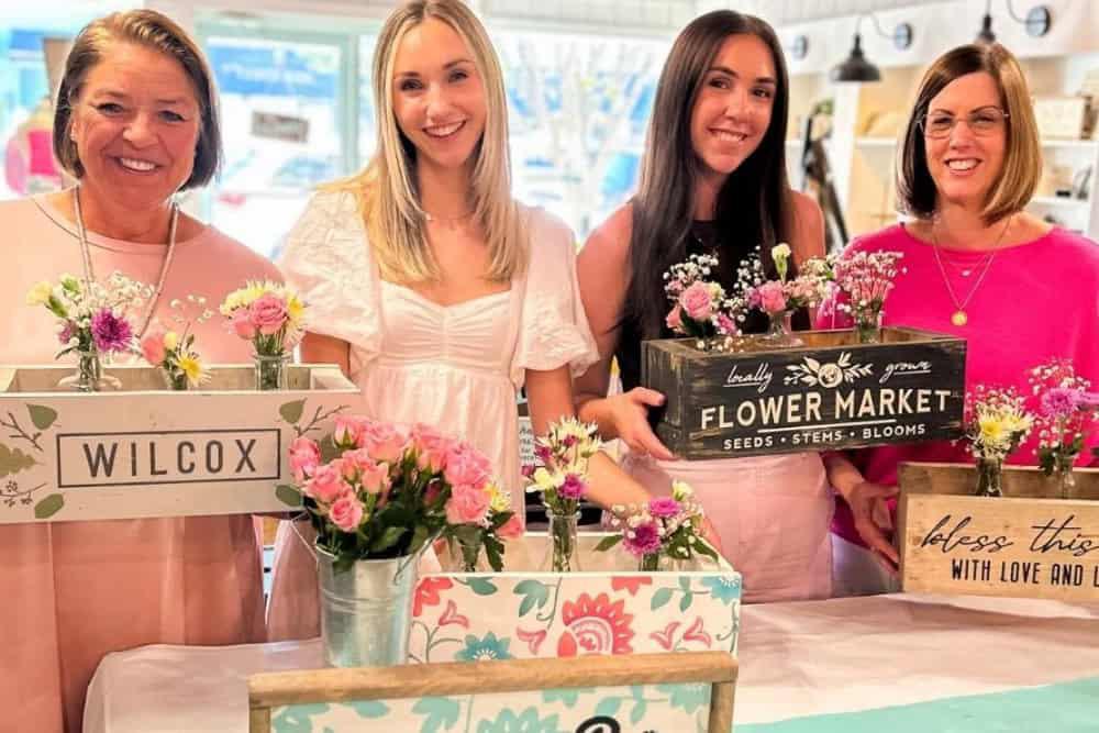 Four women smiling and holding flower arrangements in wooden boxes, each with different text and decorative designs, in a brightly lit indoor setting, showcasing one of the top 8 bachelorette party ideas in Tallahassee.